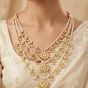 Puede incluir: Un collar de varias capas con hileras de pequeñas perlas blancas y detalles en metal dorado. El diseño incluye tres colgantes centrales en forma de flor con piedras incrustadas y racimos de perlas colgantes en cada una de las capas.