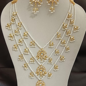 以下が含まれることがあります： 複数の白い真珠のストランドと金色の花のペンダントとアクセントが特徴のネックレスとイヤリングのセット。イヤリングは同様のデザインで、大きな花のペンダントとぶら下がっている真珠が付いています。ジュエリーはマネキンに展示されています。