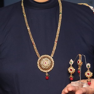 Op de afbeelding: Een gouden ketting met een ronde hanger bezet met rode en groene stenen. Passende oorbellen met rode en groene stenen zijn ook inbegrepen.