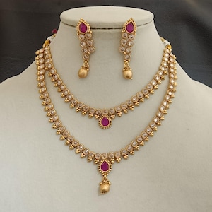 Può includere: Un set di gioielli color oro con una collana a strati e orecchini abbinati. La collana e gli orecchini sono adornati con pietre rotonde trasparenti e pietre centrali a forma di goccia di colore rubino. Il set è esposto su un manichino.