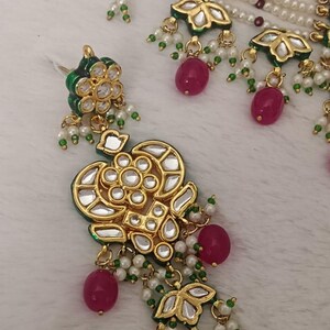 May include: Close-up of an ornate gold-colored jewelry set featuring a necklace and earrings. The set includes gold pendants with clear and green accents, red beads, and white pearls. The jewelry is set against a white, textured background.