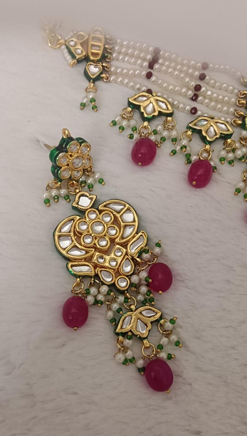 May include: Close-up of an ornate gold-colored jewelry set featuring a necklace and earrings. The set includes gold pendants with clear and green accents, red beads, and white pearls. The jewelry is set against a white, textured background.