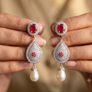 May include: A pair of drop earrings featuring an oval-cut red gemstone surrounded by clear stones, connected to a teardrop-shaped pendant encrusted with clear stones and a central red cluster, finished with a dangling white pearl drop.