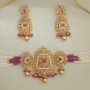 Può includere: Un set di gioielli color oro con una collana girocollo e orecchini abbinati. La collana e gli orecchini hanno un design floreale con perline rosa e bianche e pietre preziose rosa. Il set è esposto su una superficie chiara.