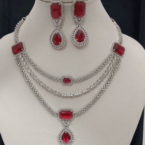Puede incluir: Un collar de plata de tres hilos con acentos de piedras preciosas rojas. El collar tiene un colgante de lágrima con una piedra preciosa roja. También se incluyen pendientes a juego con piedras preciosas rojas.