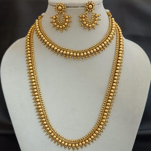 Op de afbeelding: Een goudkleurige sieradenset met een lange ketting, een kortere ketting en bijpassende oorbellen. De sieraden zijn versierd met kleine, ronde kralen en decoratieve elementen, tentoongesteld op een witte mannequinborst.
