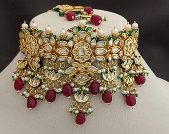 May include: A gold choker necklace and bracelet set. The necklace has floral designs with mirrored accents, green enamel, and red teardrop beads. Small white pearls and green beads add detail. The set is ornate and decorative.