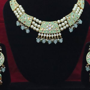 May include: A green and gold necklace and earrings set with pearl accents. The necklace has a layered design with three rows of pearls and a central pendant with a floral motif. The earrings are dangle style with a similar floral design.