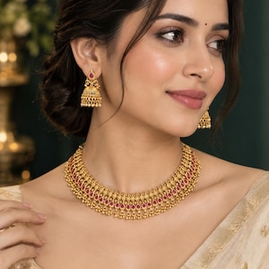 Op de afbeelding: Een goudkleurige ketting en bijpassende jhumka oorbellen set met ingewikkeld metaalwerk en kleine, druppelvormige roze stenen. De ketting heeft een gelaagd ontwerp met bungelende gouden kralen, terwijl de klokvormige oorbellen een centrale roze steen hebben.