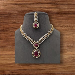 May include: A gold-toned necklace and matching pendant set with clear and ruby-colored gemstones. The necklace features a leaf-like design with a large ruby-colored teardrop pendant. The set is displayed on a gray mannequin head against a wooden background.