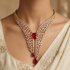May include: A gold-toned necklace featuring a central rectangular red gemstone, two dangling pear-shaped red gemstones, and numerous small, clear, marquise-cut and round-cut stones arranged in a V-shaped, leaf-like pattern across the collar.