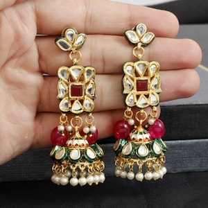 May include: A pair of gold earrings with red and green stones. The earrings feature a square design with a smaller square in the centre. The earrings hang from a chain with a small pearl at the end.