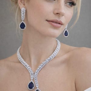 Può includere: Elegante parure di gioielli con collana e orecchini abbinati. La collana e gli orecchini hanno una montatura color argento, adornata da numerose piccole pietre trasparenti. Ogni pezzo presenta una grande gemma blu scuro a forma di goccia.