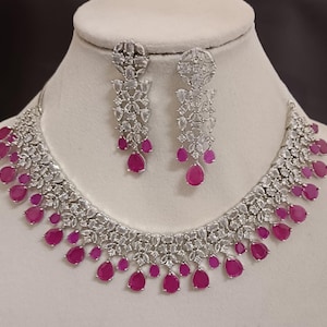 May include: A silver-toned necklace and earring set. The necklace and earrings feature a floral design with clear stones and teardrop-shaped, magenta-colored stones. The jewelry is displayed on a white mannequin bust.