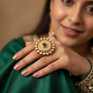 Op de afbeelding: Een goudkleurige ring met een centrale, ronde, donkerrode steen, omringd door een cirkel van kleine, heldere stenen. De ring heeft een bloemenontwerp en staat tegen een groene stoffen achtergrond.