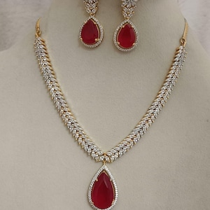Puede incluir: Un conjunto de joyas de tono dorado con un collar y pendientes a juego. El collar y los pendientes tienen una piedra preciosa central en forma de lágrima, de color rojo intenso, rodeada por un halo de pequeñas piedras transparentes. El collar tiene un diseño de hojas.