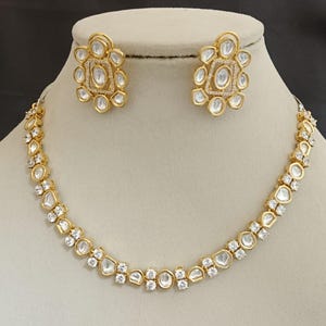 May include: A gold-coloured jewellery set comprising a necklace and matching earrings. The necklace and earrings are decorated with clear, sparkling gemstones and a gold setting. The earrings have a floral design, and the necklace has a delicate, symmetrical pattern.