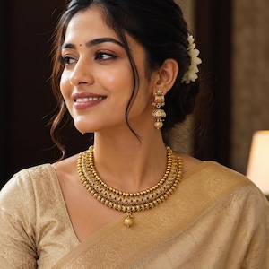 Peut inclure: Un collier en or composé de deux rangées superposées de perles texturées avec de petits accents rouges et une perle centrale suspendue, assorti de boucles d'oreilles jhumka en or en forme de cloche avec une petite perle pendante.