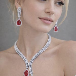 May include: Elegant jewelry set featuring a necklace and matching earrings. The necklace and earrings are adorned with sparkling white stones and teardrop-shaped red gemstones. The jewelry is designed to be worn for special occasions.