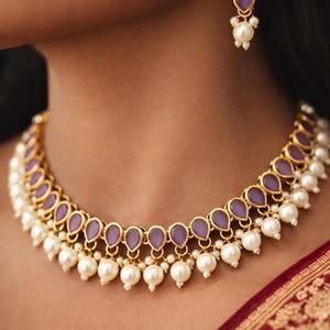 Puede incluir: Un conjunto de collar y pendientes dorados a juego. El collar presenta piedras moradas en forma de lágrima y perlas blancas. Los pendientes tienen tres piedras moradas y una perla. La joyería se muestra en una persona.