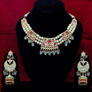 May include: A white and gold necklace with green and red floral accents and a matching pair of earrings. The necklace is made of pearls and has a layered design. The earrings are crescent shaped and have a dangling bell charm.
