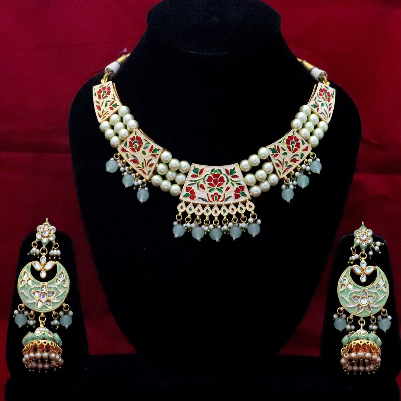May include: A white and gold necklace with green and red floral accents and a matching pair of earrings. The necklace is made of pearls and has a layered design. The earrings are crescent shaped and have a dangling bell charm.
