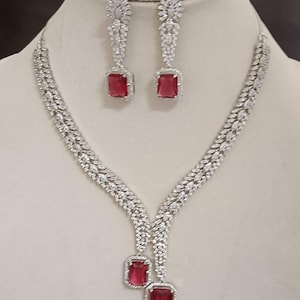 Puede incluir: Un conjunto de joyas plateadas con un collar y pendientes a juego. El collar y los pendientes están adornados con piedras claras y brillantes y presentan piedras centrales rectangulares de color rojo rubí. Los pendientes son de estilo colgante y el collar tiene un colgante en forma de Y.