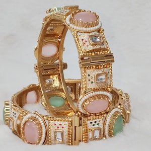 May include: Two gold-colored bangles with intricate designs. Each bangle features oval-shaped pink and green gemstones, surrounded by white and gold detailing. The bangles have a hinged clasp and are decorated with small, square, clear stones.