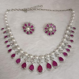 Pode incluir: Um conjunto de joias prateadas com colar e brincos a condizer. O colar e os brincos são adornados com pérolas brancas e pedras preciosas vibrantes em forma de gota, cor de rubi. Os brincos têm um design floral com uma pérola central.