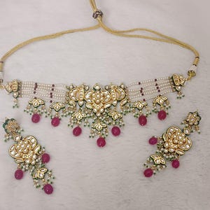 May include: A gold-toned necklace and matching earrings set. The necklace features a choker style with multiple strands of white beads, gold filigree, and dangling red and green accents. The earrings mirror the necklace design.
