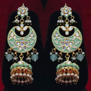 May include: A pair of green and gold earrings with intricate floral designs and pearl accents. The earrings feature a crescent moon shape with a dangling jhumka style bell.