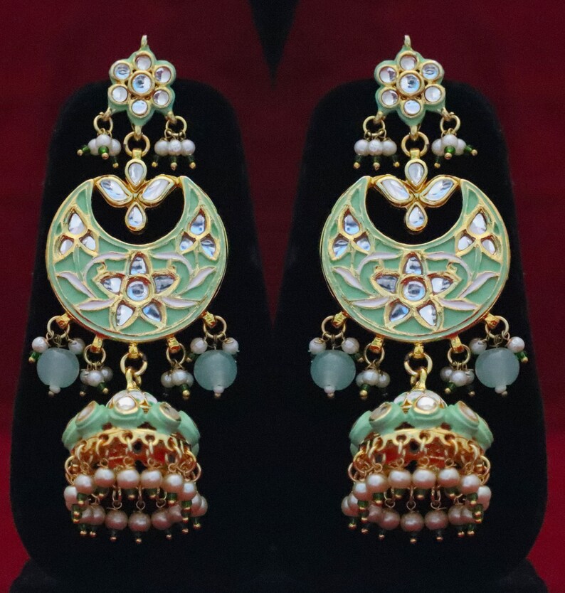 May include: A pair of green and gold earrings with intricate floral designs and pearl accents. The earrings feature a crescent moon shape with a dangling jhumka style bell.