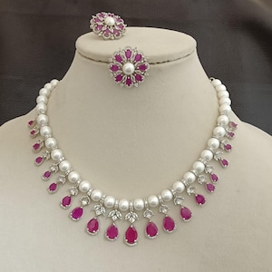 May include: A pearl necklace and ring set displayed on a mannequin. The necklace has large white pearls with ruby-coloured teardrop pendants. The ring features a central pearl surrounded by ruby-coloured stones. The jewellery is shown on a cream-coloured bust.
