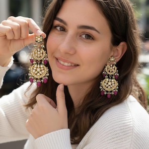 May include: Gold chandelier earrings with detailed design. The earrings have multiple circular tiers with white gemstones and small red beads. Each earring is topped with a floral design.