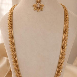 May include: A gold-toned jewelry set consisting of a long necklace, a pair of drop earrings, and a maang tikka. Each piece features a repeating leaf-shaped motif with intricate textured detailing and small beaded accents along the edges.