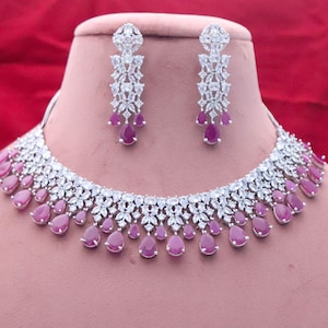 May include: A silver necklace with a row of teardrop-shaped pink stones and a matching pair of earrings. The necklace and earrings are set with clear cubic zirconia stones.