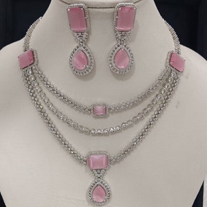Może przedstawiać: Trójrzędowy srebrny naszyjnik z różowymi kamieniami i pasującą parą kolczyków. Naszyjnik ma wisiorek w kształcie łzy z różowym kamieniem.