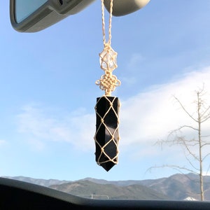 May include: A black obsidian crystal point wrapped in macrame hanging from a rearview mirror in a car. The crystal is a dark black color and is surrounded by a beige macrame net. The crystal is hanging from a beige cord.