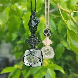May include: Two crystal pendants hanging from black and white macrame cords. The crystals are clear and faceted. The pendants are hanging from a branch with green leaves.