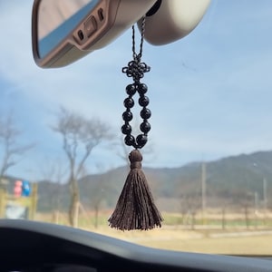May include: A black beaded car charm with a brown tassel hanging from the rear view mirror of a car. The charm is made of black beads and has a brown tassel at the bottom.