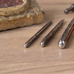 Op de afbeelding: Drie metalen gereedschappen met afgeronde uiteinden, gelegd op een houten oppervlak. De gereedschappen zijn van verschillende lengtes en hebben een getextureerde afwerking.