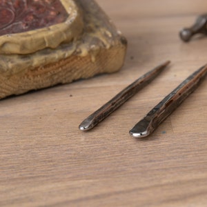 Könnte beinhalten: Zwei Metallwerkzeuge mit abgerundeten Spitzen, die auf einer Holzoberfläche liegen. Die Werkzeuge werden zum Formen von Metall verwendet.