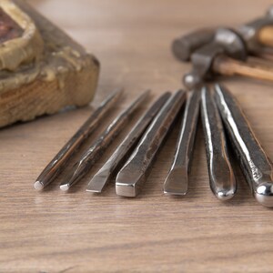 Op de afbeelding: Een set van acht metalen gereedschappen voor het maken van sieraden, waaronder een hamer en verschillende beitels, gerangschikt op een houten oppervlak.