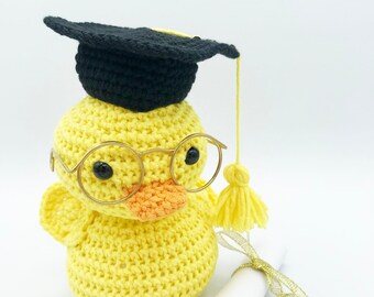 May include: A yellow crocheted duck wearing a black graduation cap and gold-rimmed glasses, holding a rolled-up diploma with a gold ribbon.