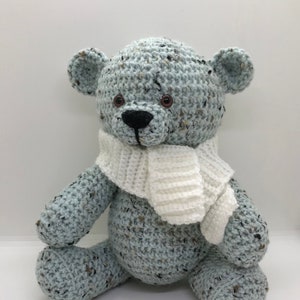 May include: A crocheted blue teddy bear wearing a white crocheted scarf. The bear is sitting on a white surface.