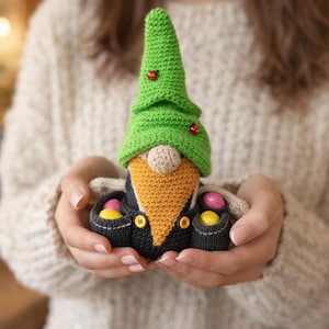 May include: A crocheted gnome figurine featuring a tall green hat adorned with two small red beads, a tan nose, a long mustard-yellow beard, and a black outfit with yellow buttons. The gnome holds two small colorful eggs in its pockets.