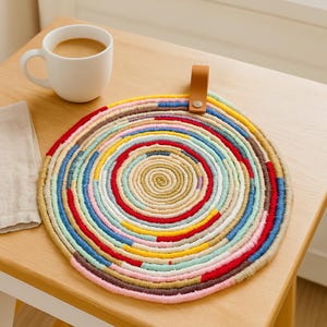 Puede incluir: Un salvamanteles circular multicolor hecho de cuerda trenzada en espiral. El salvamanteles presenta una variedad de colores, incluyendo rojo, azul, amarillo y beige. Tiene un pequeño lazo de cuero marrón para colgar. Una taza blanca con café está cerca.