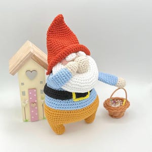 Może przedstawiać: Puchaty krasnal z czerwoną czapką, niebieską koszulą i pomarańczowymi spodniami trzyma mały wiklinowy koszyk. Krasnal stoi przed małym drewnianym domkiem.