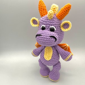 May include: A purple crocheted dragon with orange and yellow accents. The dragon has a friendly expression and is standing on a white background.