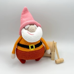 May include: A crocheted gnome doll with a pink hat, white beard, and orange pants. The gnome is standing next to a wooden toy mallet and a wooden block.